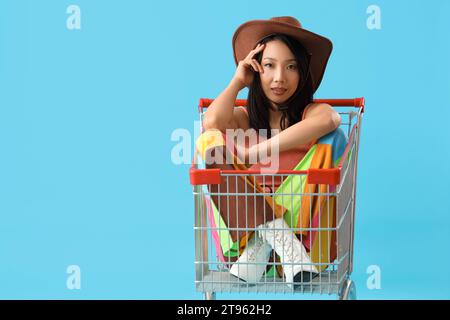 Schöne junge, stilvolle asiatische Frau, die im Einkaufswagen auf blauem Hintergrund sitzt Stockfoto