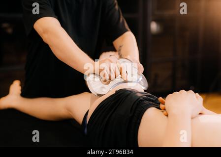 Therapeutin mit IASTM-Instrumentenbehandlung, Mann erhält Weichteilbehandlung am Bein mit guasha-Edelstahlwerkzeug, Osteopathie Stockfoto