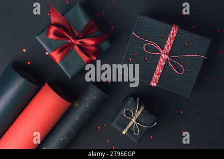 Geschenkboxen Verpackung mit roter Schleife und Rollen Geschenkpapier auf schwarzem Hintergrund. Festliche Weihnachten, Neujahrsfeierlichkeiten. Draufsicht Stockfoto