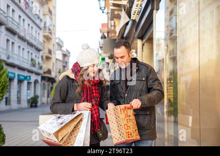 Lächelndes junges hispanisches Paar in warmen Kleidern, das auf der Straße der Stadt nach unten blickt, und männlich, der am Tag gekaufte Artikel in der Tasche einer Frau in der Mütze zeigt Stockfoto