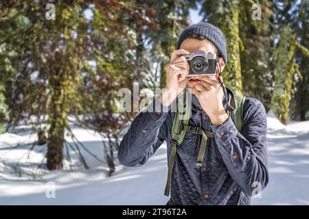 Porträt eines männlichen Reisenden in warmer Kleidung und Rucksack, der Fotos mit einer Fotokamera macht, während er auf verschneiten Waldgebieten im Yosemite California steht Stockfoto