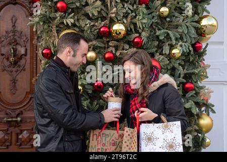 Seitenansicht des jungen hispanischen Paares mit Taschen, die nach unten schauen, während sie neben einem geschmückten Weihnachtsbaum mit Kugeln stehen und Süßigkeiten bei Tageslicht teilen Stockfoto