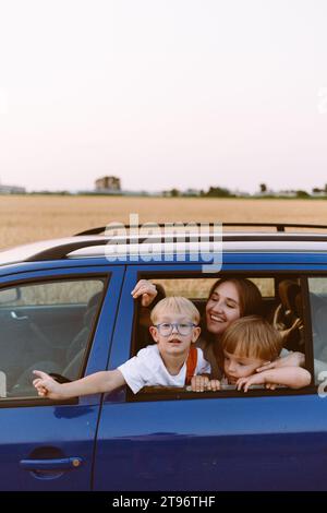 Fröhliche junge Mutter mit entzückenden, neugierigen kleinen Söhnen, die auf der Rückbank auf blauem Auto sitzen und in der Nähe des Weizenfeldes auf dem Land lächeln Stockfoto