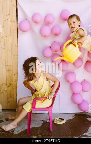 Mädchen sitzt auf einem Stuhl neben der Wand mit pinkfarbenen Ballons, während der kleine Bruder in den Händen von Eltern sitzt, die vorgeben, reines Wasser aus der Dose zu geben Stockfoto