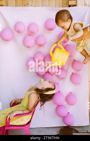 Mädchen mit geöffnetem Mund, das auf einem Stuhl neben der Wand mit rosa Ballons sitzt, während der kleine Bruder in den Händen einer Mutter, die vorgibt, reines Wasser aus der Dose zu geben Stockfoto