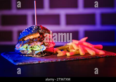 Leckerer Burger gegrillt zwischen Brötchen in der Nähe mit Chips Scheiben auf beleuchtetem dunklem Hintergrund Stockfoto