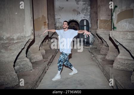 Ein ganzer Körper erfahrener Männer in Freizeitkleidung, der in die Kamera blickt, während er Breakdance-Bewegungen in einem alten verlassenen Gebäude vorführt Stockfoto