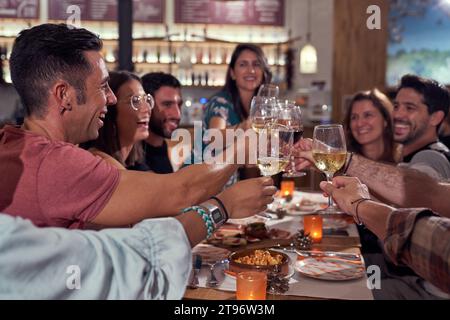 Gruppe lächelnder junger Freunde, die an einem Tisch in der Bar sitzen und mit Weingläsern anstoßen, während sie gemeinsam den Anlass feiern Stockfoto