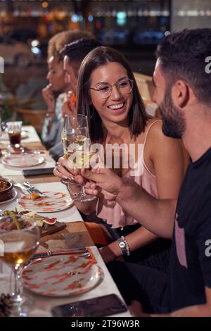 Glücklicher junger Mann und verliebte Frau aus ethnischer Herkunft, die am Holztisch mit Gläsern Wein sitzen und beim Abendessen in der Bar am Abend toasten Stockfoto