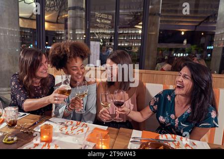 Gruppe glücklicher, multirassischer Freundinnen, die während der festlichen Veranstaltung im modernen Restaurant zusammen an einem Serviertisch sitzen Stockfoto