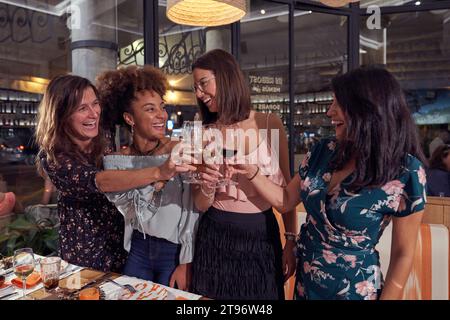 Eine Gruppe positiver, multirassischer Freundinnen, die Toast vorschlagen, während sie Weingläser anschlagen und sich während der festlichen Veranstaltung im Restaurant anschauen Stockfoto