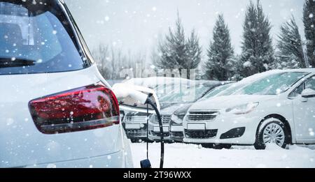 Nahaufnahme eines Elektroautos mit angeschlossenem Ladekabel auf dem verschwommenen Hintergrund eines schneebedeckten geparkten Autos. Stockfoto