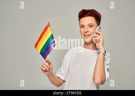 Glückliche rothaarige nicht-binäre Person, die eine kleine LGBT-Flagge hält und auf dem Handy auf Grau spricht Stockfoto