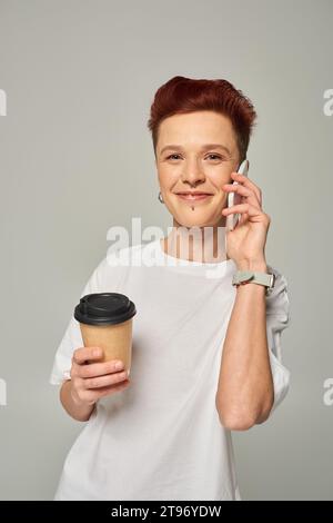 Lächelnde rothaarige, queere Person, die Kaffee hält, um in einer Pappbecher zu gehen und auf dem Smartphone auf Grau zu sprechen Stockfoto