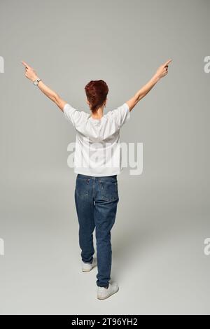 Rückansicht einer rothaarigen queeren Person in weißem T-Shirt und Jeans, die mit den Fingern auf Grau zur Seite zeigt Stockfoto