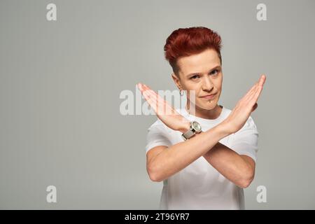 Strenge und ernsthafte Bigender-Person mit rotem Haar, die Stop-Geste zeigt und die Kamera auf Grau betrachtet Stockfoto