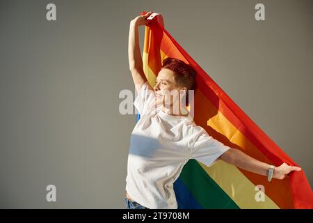 Glückliche und einzigartige queere Person in weißem T-Shirt, die mit LGBT-Flagge in Regenbogenfarben auf grauem Hintergrund posiert Stockfoto