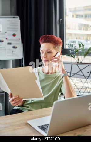 Queere Person, die mit dem Smartphone spricht und Dokumente in der Nähe eines Laptops in einem modernen Büro ansieht Stockfoto