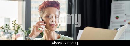 Queere Person, die auf dem Smartphone spricht und Dokumente in einem modernen Büro ansieht, horizontales Banner Stockfoto