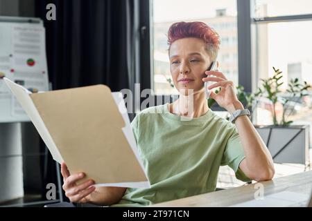 Queere Person, die mit dem Smartphone spricht und Dokumente im modernen Büro ansieht, Multitasking Stockfoto