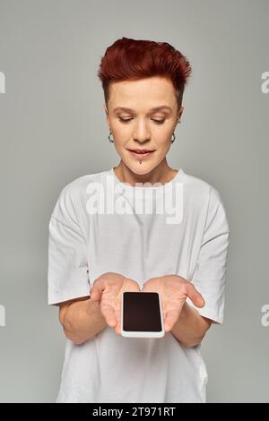 Rothaarige nicht-binäre Person in weißem T-Shirt, die Handy mit leerem Bildschirm auf grauem Hintergrund hält Stockfoto