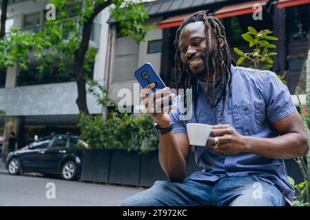 Junger afrikanischer Mann mit Dreadlocks, glücklich lächelnd und genießt den Tag, wenn er Kaffee trinkt und draußen telefoniert, soziale Netzwerke überprüft und benachrichtigt Stockfoto