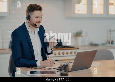 Fröhlicher Geschäftsmann in einem Headset mit Wasser nimmt an einem virtuellen Meeting von einem hellen Heimbüro Teil Stockfoto