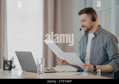 Ein fokussierter Mann mit Headset prüft Dokumente, während er Remote arbeitet, und zeigt Produktivität und Konzentration in der häuslichen Umgebung Stockfoto