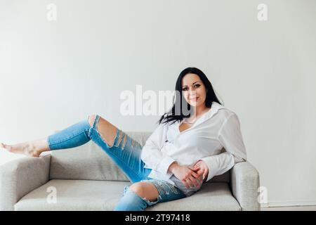 Schöne brünette Frau, die auf grauem Sofa in weißem Zimmer sitzt Stockfoto