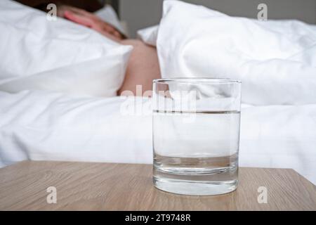 Glas mit Wasser auf Holztisch und Schlafperson in einem Zimmer Stockfoto