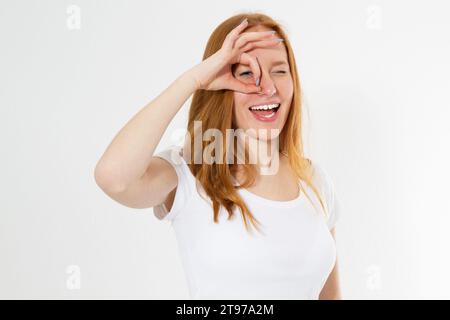 Rotes Kopfmädchen in farbenfroher Kleidung hält ihre Finger um den Kopierraum der Augen isoliert, junge Frau zeigt Okays und schaut durch Stockfoto