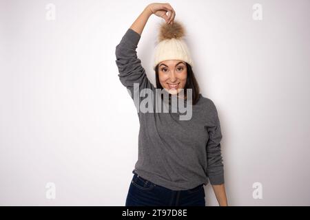 Gut aussehende brünette junge Frau hält Hand nah am Gesicht, lächelt angenehm, trägt Hut und grauen Strickpullover, genießt angenehmen Moment, drückt gut aus Stockfoto