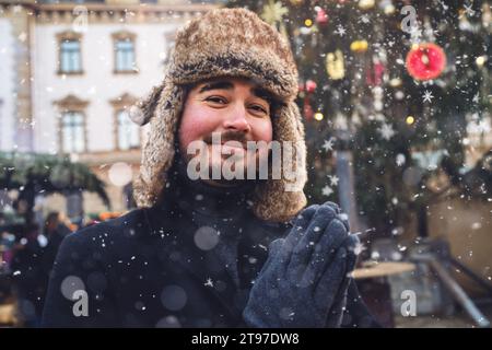 Mann mit Pelzhut und Wintermantel lächelt sanft, Schneeflocken fallen um ihn herum, Weihnachtsmarkt Lichter im Hintergrund. Stockfoto
