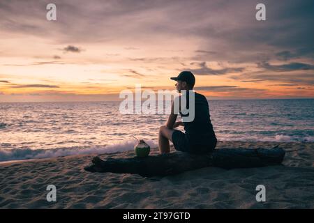 Mann sitzt am tropischen Sandstrand mit Kokosnussgetränk und genießt den farbenfrohen Sonnenuntergang über dem Meer. Themen für Alleinreisende und Buget-Reisen. Stockfoto