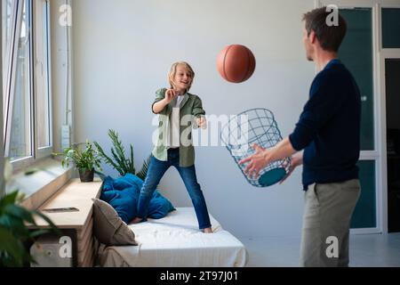 Glücklicher Junge, der Basketball wirft, während Vater zu Hause einen Papierkorb hält Stockfoto