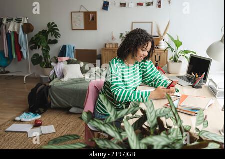 Glückliches Mädchen im Teenageralter, das das Smartphone am Schreibtisch in einem schmutzigen Schlafzimmer benutzt Stockfoto