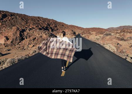 Junge Frau, die mit Decke in der Nähe der vulkanischen Landschaft an sonnigem Tag spaziert Stockfoto