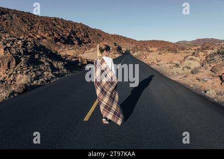 Junge Frau mit Decke, die auf der Straße in der Nähe vulkanischer Landschaften läuft Stockfoto