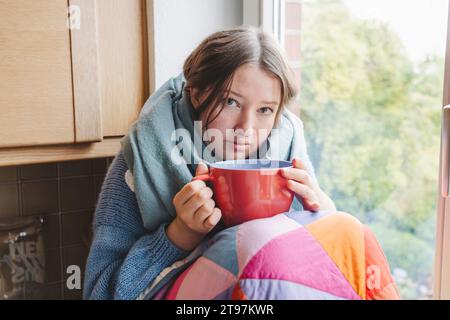 Das kranke Mädchen, in eine Decke gewickelt und zu Hause mit einer Kaffeetasse gesessen Stockfoto