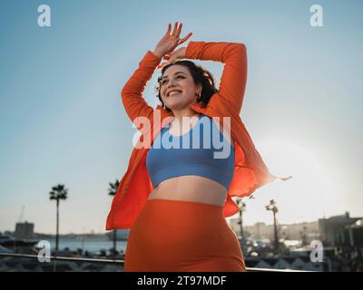 Glückliche Frau in Sportbekleidung, die mit erhobenen Armen unter dem Himmel steht Stockfoto