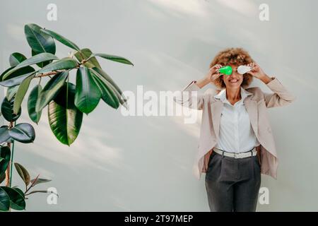 Lächelnde Geschäftsfrau, die Glühbirnen über den Augen vor der Wand hält Stockfoto