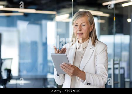 Die leitende Geschäftsfrau, Direktorin und Gründerin des Unternehmens steht im Büro und führt auf einem Tablet ein Videogespräch mit Partnern und Kunden. Gesten mit Händen, erklärt, bespricht. Stockfoto