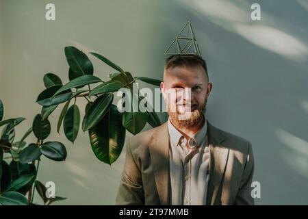 Lächelnder Geschäftsmann, der eine Metallpyramide auf dem Kopf vor der Wand balanciert Stockfoto