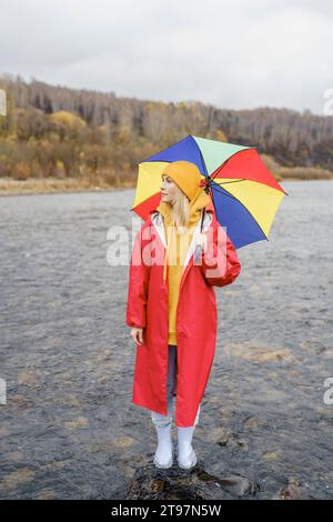 Kontemplative Frau mit rotem Regenmantel, die mit Regenschirm im Flusswasser steht Stockfoto