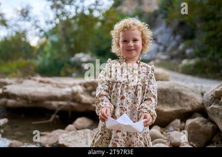 Lächelndes, süßes Mädchen, das ein Papierboot vor Felsen hält Stockfoto