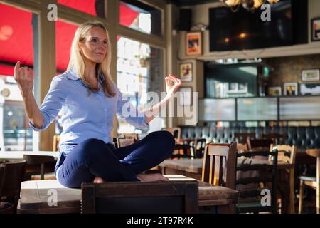 Blonde Geschäftsfrau, die im Café auf dem Tisch sitzt und Yoga macht Stockfoto