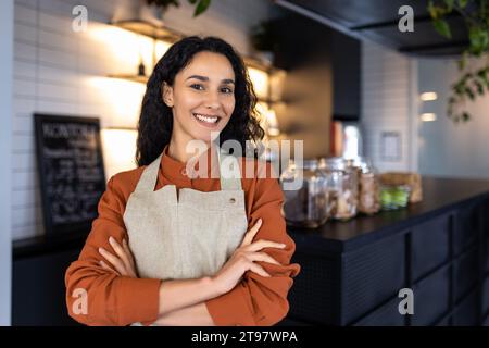 Porträt eines jungen, fröhlichen, erfolgreichen lateinamerikanischen Kleinunternehmers, schlanker Chef, der lächelnd und mit überkreuzten Armen in die Kamera blickt, Geschäftsfrau am Eingang des Cafés Restaurant, einlädt Besucher. Stockfoto