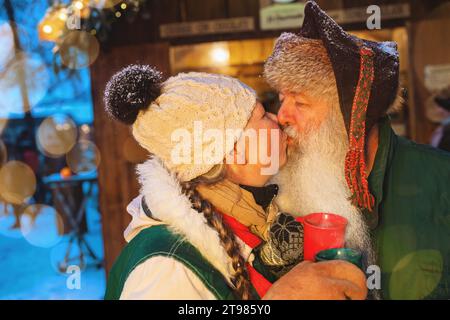 Seniorenpaar küsst sich auf dem Weihnachtsmarkt, hält rote Tassen, Bokeh-Lichter im Hintergrund Stockfoto