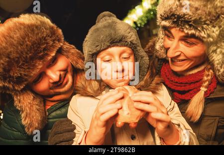 Familie genießt Essen auf einem Weihnachtsmarkt, Kind isst ein Wurstsandwich mit Freude Stockfoto