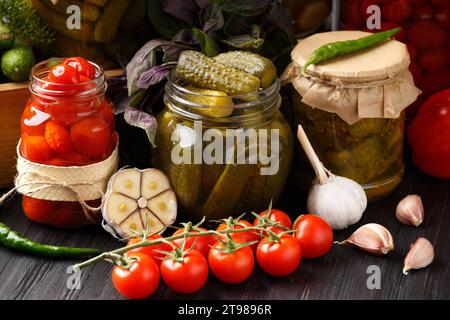 Konservierte Kirschtomaten und Gurken in Gläsern, frisches Gemüse, Gewürze und Kräuter für Marinade auf hölzernem Hintergrund. Eingelegtes Gemüse. Stockfoto